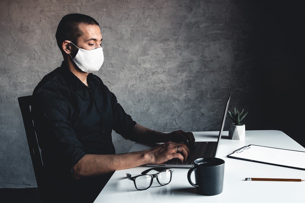 A Masked Man Works At A Computer. Pandemic, Coronavirus, Epidemic